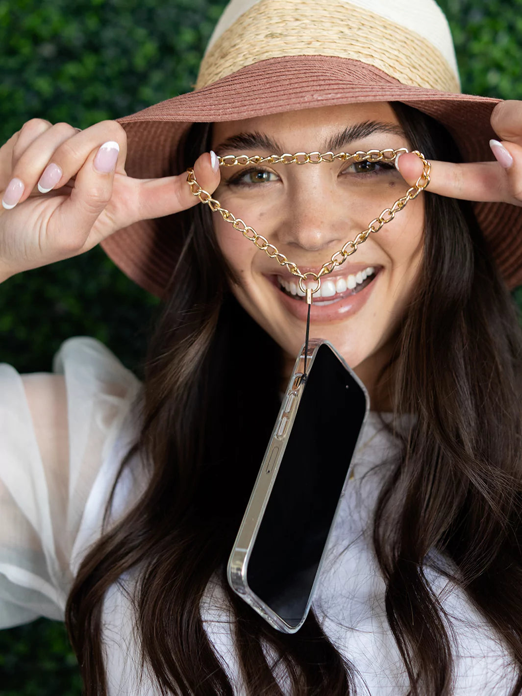 Expanding gold phone wristlet charm using 2 fingers connected to iPhone wearing hat and white blouse with green background