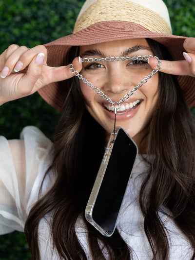 Expanding silver phone wristlet charm using 2 fingers connected to iPhone wearing hat and white blouse with green background