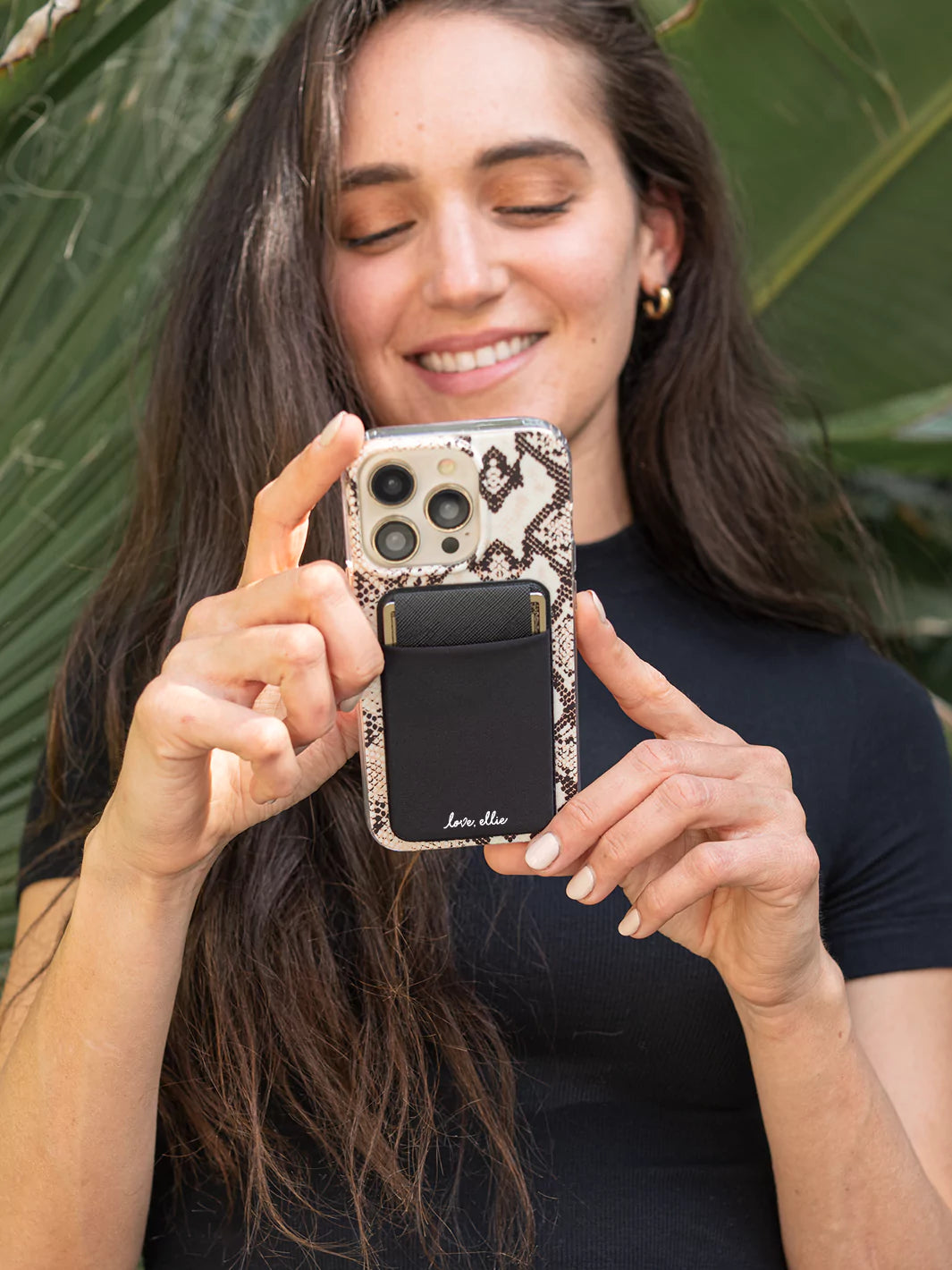 Black Stick on Phone Pocket attached to iPhone wearing black top and green plants as background