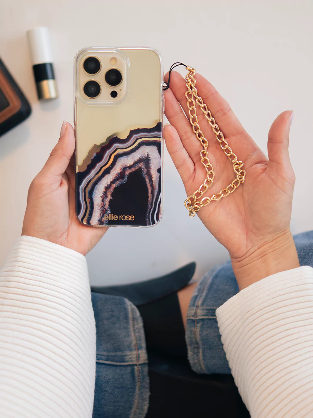 Showing gold phone wristlet charm and iPhone with black agate wearing blue jeans and white top with white background