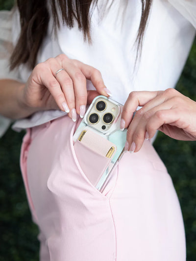 Holding a iPhone with stick on phone pocket blush attached at the back wearing white tops and blush pants