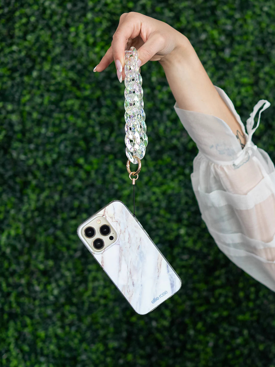 Holding iPhone latched with holographic phone wristlet charm wearing white tops and hat with green background wallpaper