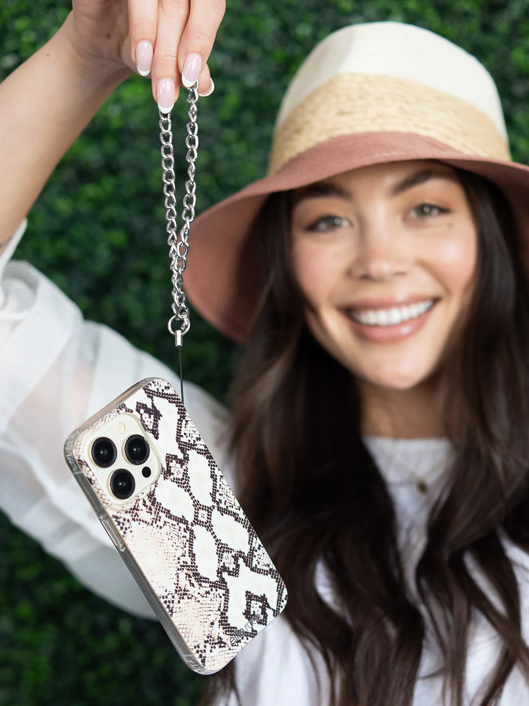 Showing gunmetal phone wristlet charm connected to iPhone with snakeskin case wearing colored hat and white top