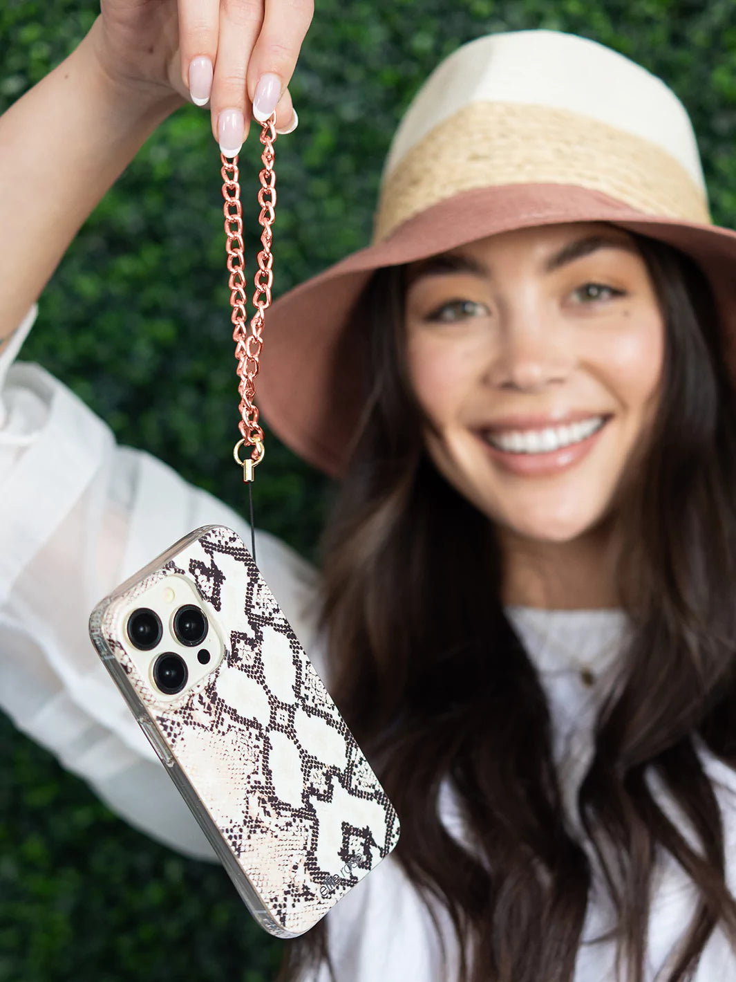 Holding Rose gold wristlet  charm latched to iphone with snakeskin case wearing hat and white dress