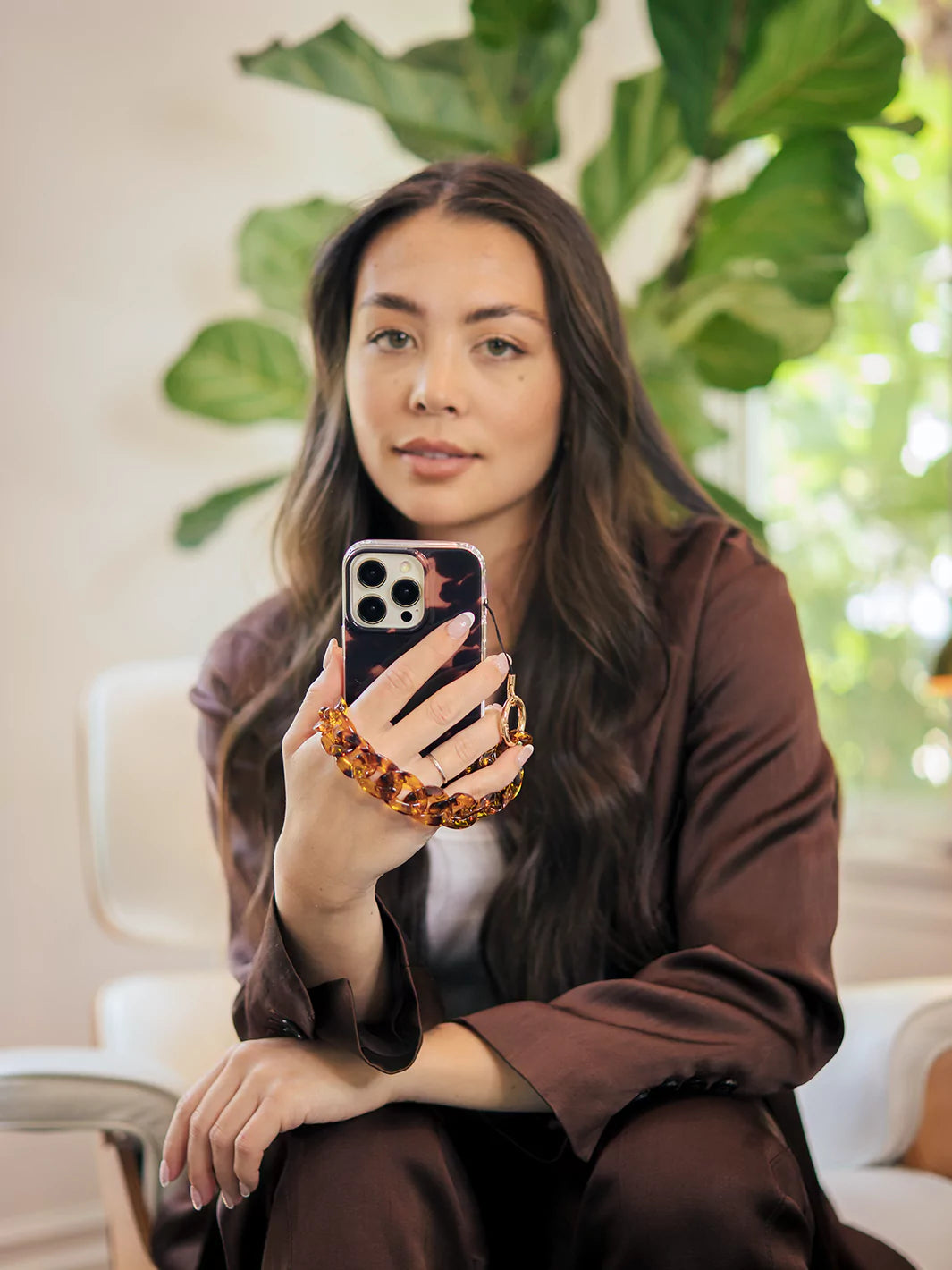 Holding and taking a picture with tortoiseshell phone wristlet charm and iphone with a background of a trees  wearing brown top and brown jeans